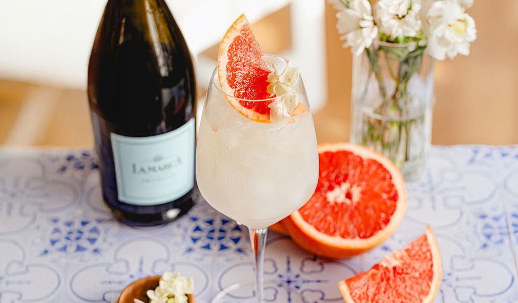 A crisp La Marca Prosecco cocktail in a wine glass over ice, garnished with a vibrant half-wheel of orange and a white floral sprig. The glass sits on a blue and white Mediterranean-style tiled surface next to a whole orange cut in half, loose orange slices, and a bottle of La Marca Prosecco.