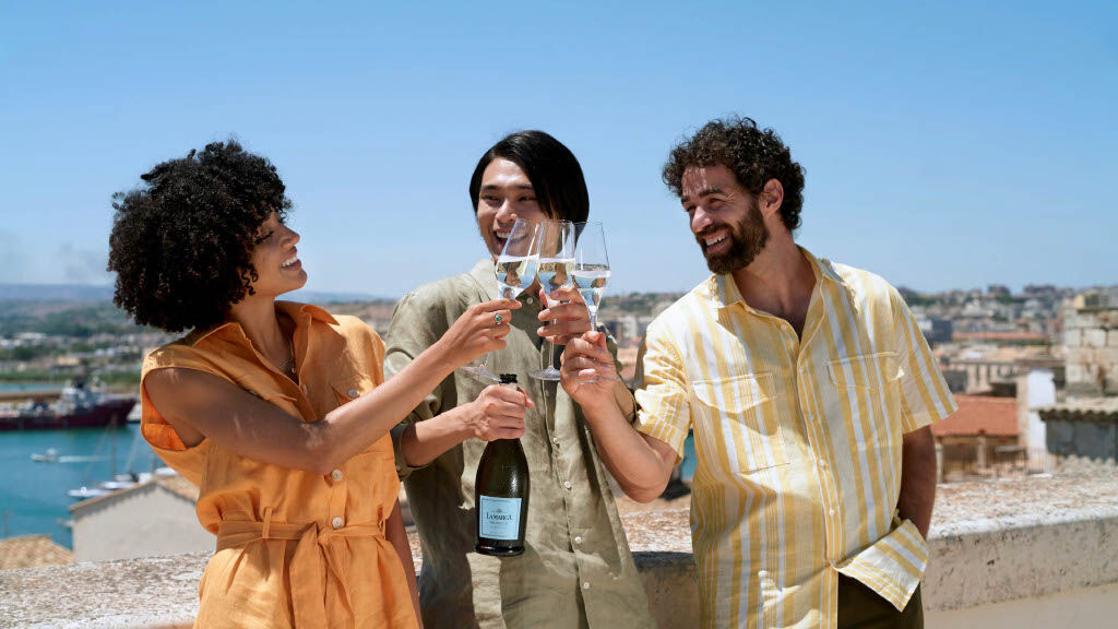 Three friends—a woman in an orange linen dress and two men in summer shirts—laugh and toast with glasses of sparkling wine on a sun-drenched rooftop overlooking a coastal harbor and city skyline, with one man holding a bottle of La Marca Prosecco.